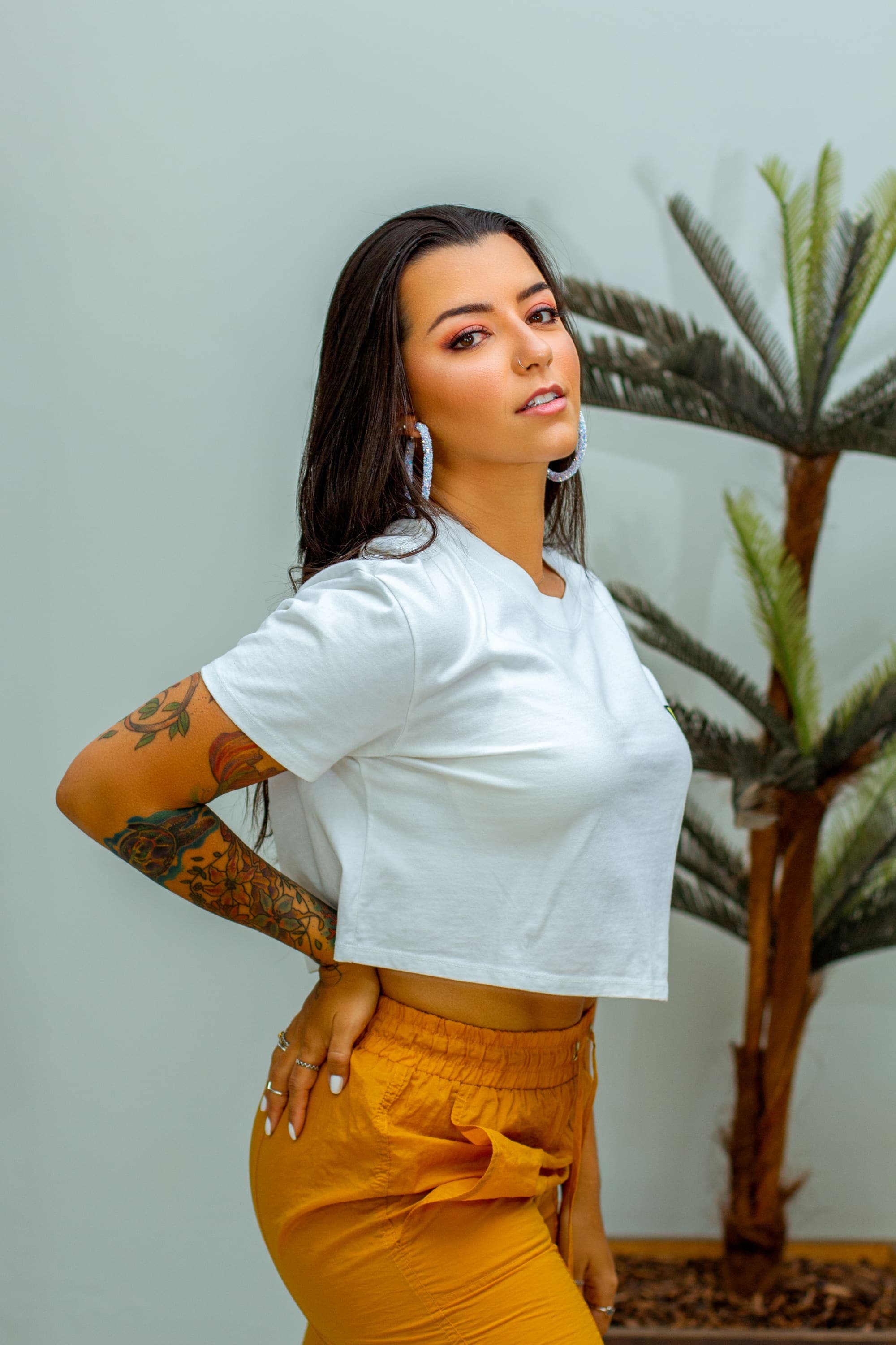 Retrato em estúdio com planta ao fundo, mulher de camiseta branca e calça caramelo, mão na cintura, em Brasília