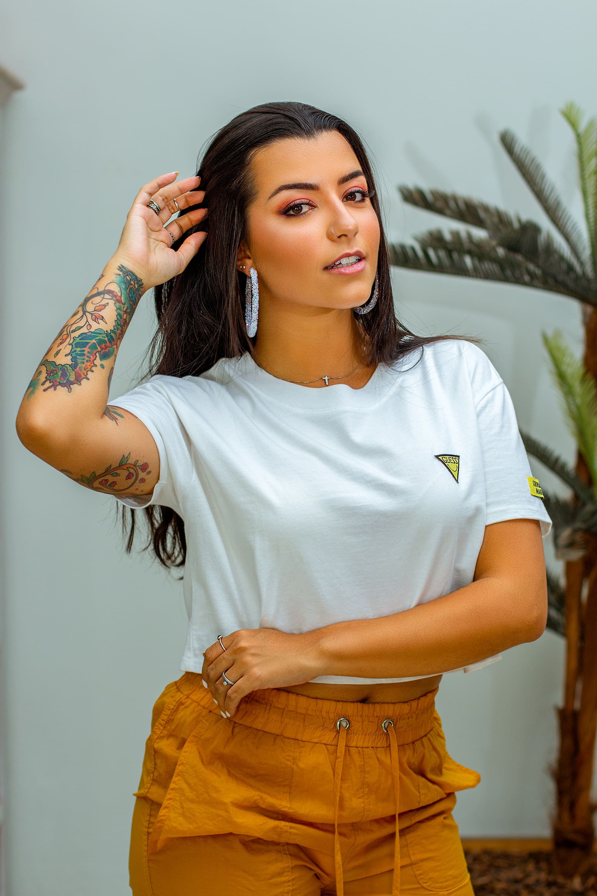 Retrato em estúdio com planta ao fundo, mulher de camiseta branca e calça caramelo, mão no cabelo, em Brasília
